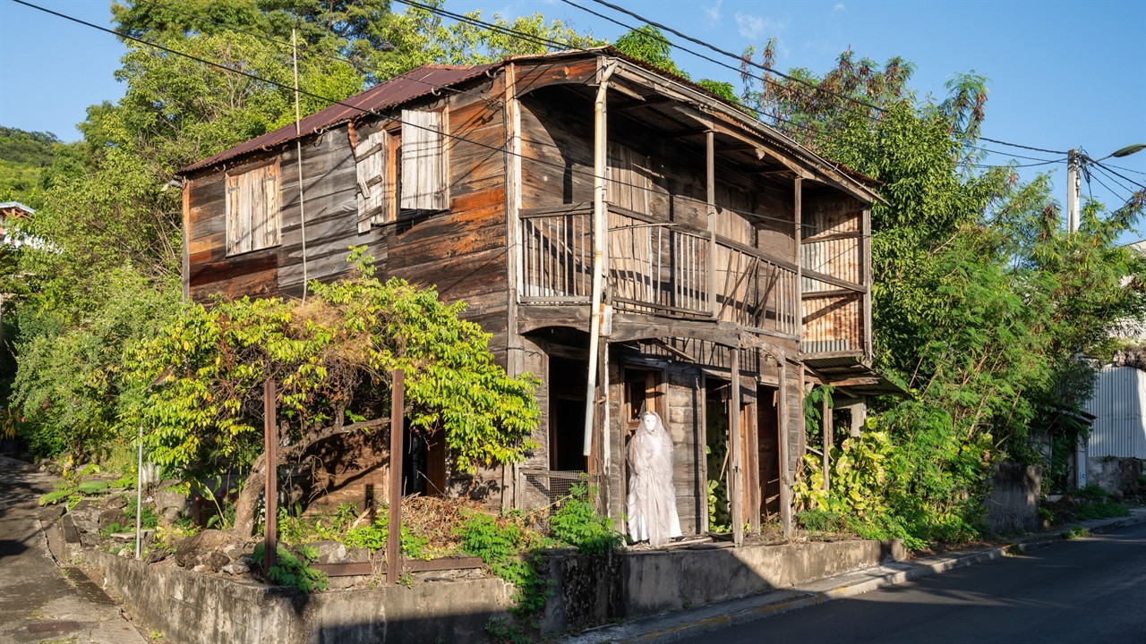 A veiled figure stands outside an old wooden house